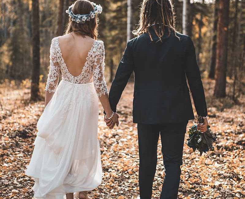 Ein modernes Brautpaar läuft durch den Wald man sieht beide von hinten,Braut mit weißem Kleid,Bräutigam im Anzug mit Rastalocken