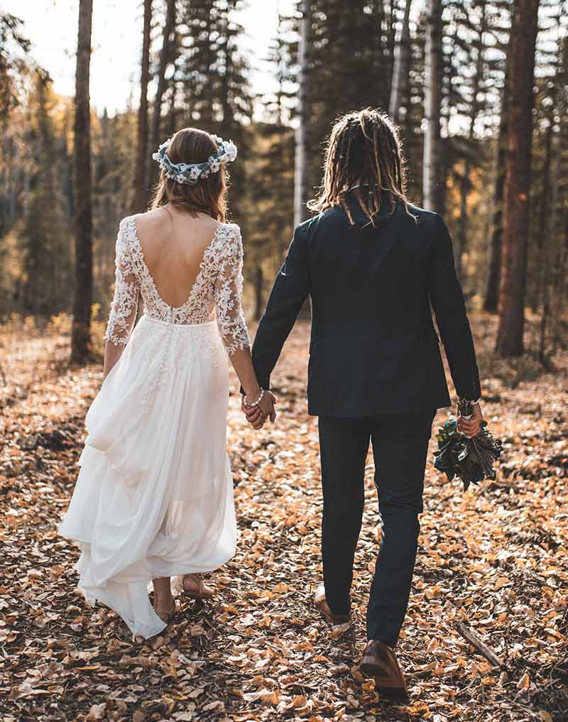 Ein modernes Brautpaar läuft durch den Wald man sieht beide von hinten,Braut mit weißem Kleid,Bräutigam im Anzug mit Rastalocken