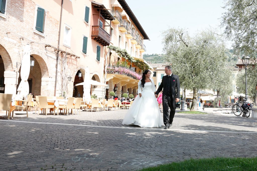 Brautpaar am Gardasee läuft entlang einer Promenade, weißes Hochzeitskleid, schwarzer Anzug, Olivenbäume und italienisches Flair
