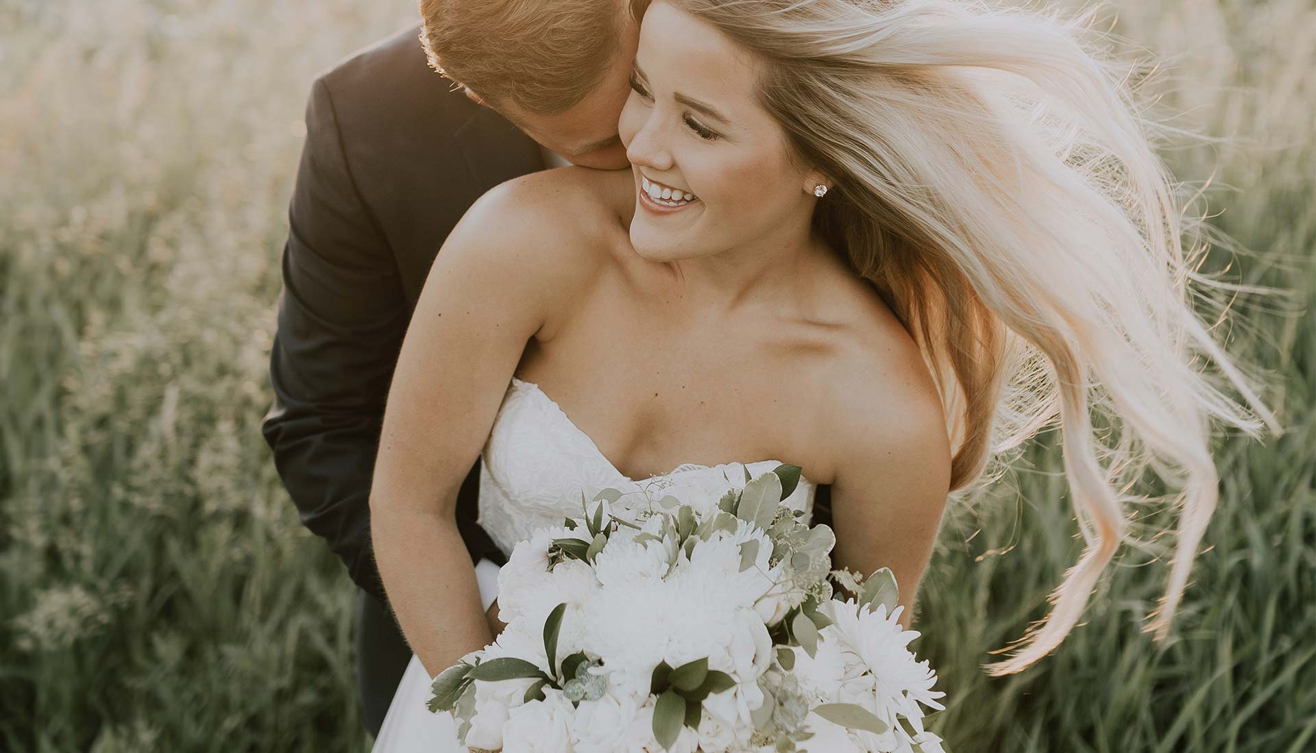 Bild einer Braut mit wehenden Haaren und weißem Hochzeitskleid, ihr Bräutigam setht hinter ihr und küsst ihren Hals